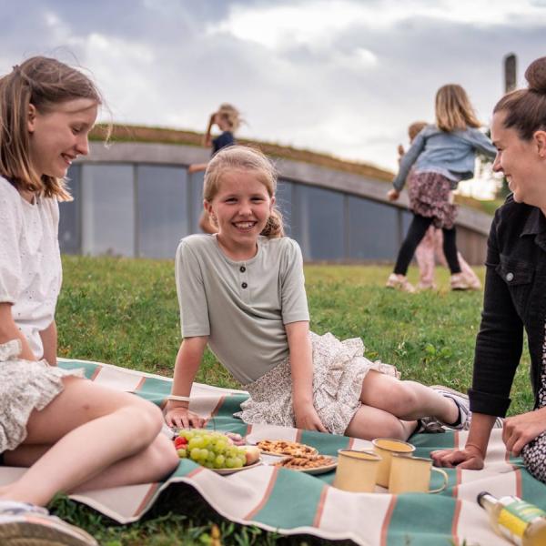 En mor og hendes to døtre sidder på et tæppe og spiser snacks. Bagved ligger besøgscenteret, bygget ind i en jordvold.