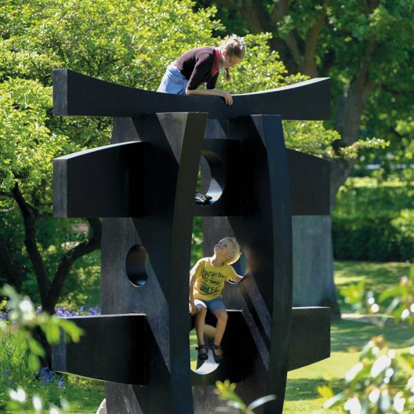 To børn leger på den store sorte skulptur "Pagode" af Hans August Andersen i Geografisk Have. Skulpturen er omgivet af frodig natur og grønne træer. 