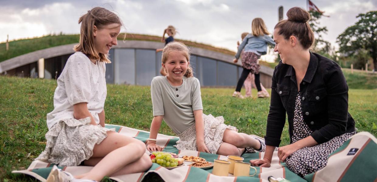 En mor og hendes to døtre sidder på et tæppe og spiser snacks. Bagved ligger besøgscenteret, bygget ind i en jordvold.