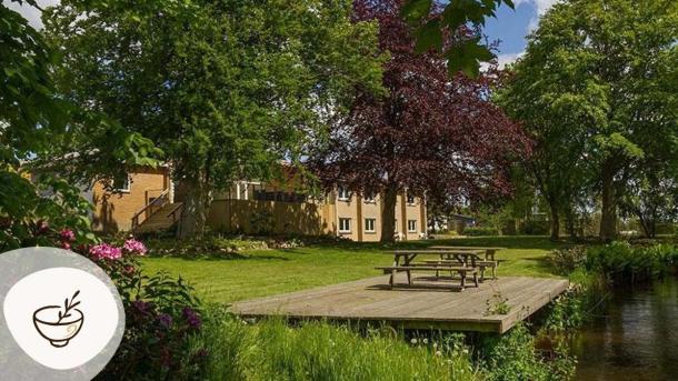 Omme Å, have og terrasse med borde-bænke-sæt bagved Sdr. Omme Kro, omgivet af grønne træer og rolige omgivelser. Kroen er en del af Gastroruten i Small Wonders