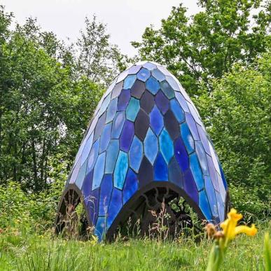 Skulpturen Pentagonia i Skulpturpark Billund, en blå mosaikformet installation med udskårne metaldetaljer ved basen. Gule påskeliljer foran skulpturen. En del af Kunstruten i Small Wonders – Heart of Jutland.