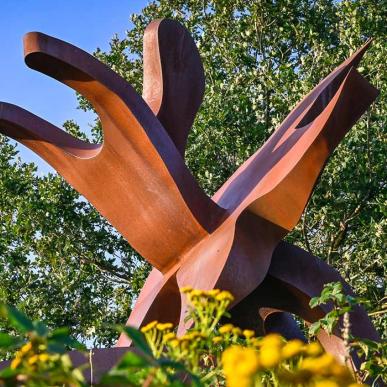 Skulpturen STJERNEDYRET i Skulpturpark Billund, set nedefra mellem grønne planter. og gule blomster. En del af Kunstruten i Small Wonders – Heart of Jutland.